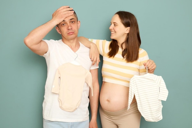 Une jeune famille mariée enceinte choisissant des vêtements pour le futur bébé, un mari fatigué, un futur père faisant un geste de paume sur le visage, isolé sur un fond vert clair.