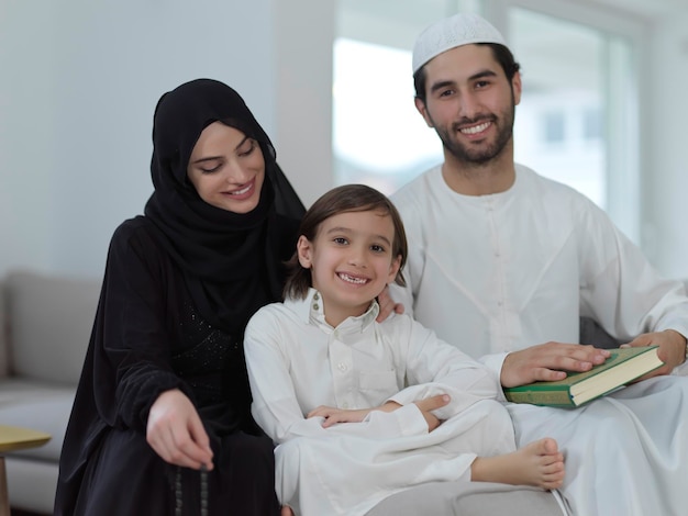 Jeune famille musulmane lisant le Coran pendant le Ramadan. Parents et fils adorant Dieu, en vêtements islamiques à la maison moderne