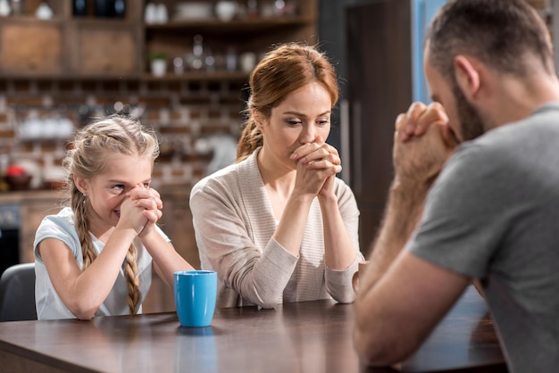 Jeune famille priant assis à la table dans la cuisine