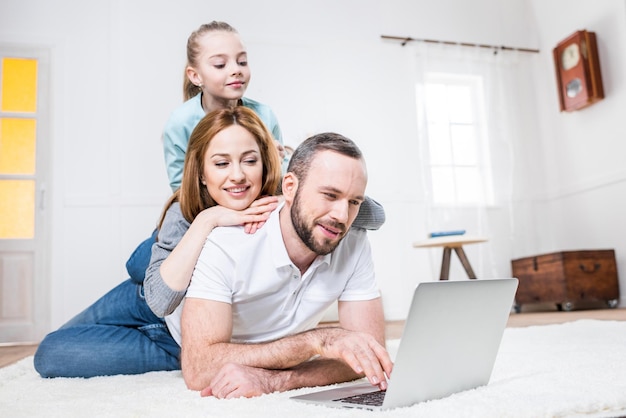 Jeune famille de trois personnes utilisant un ordinateur portable allongé sur un tapis à la maison