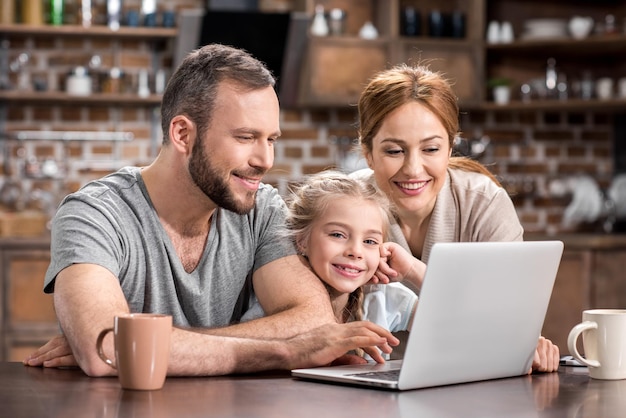 Jeune famille utilisant un ordinateur portable dans la cuisine et buvant du thé ensemble