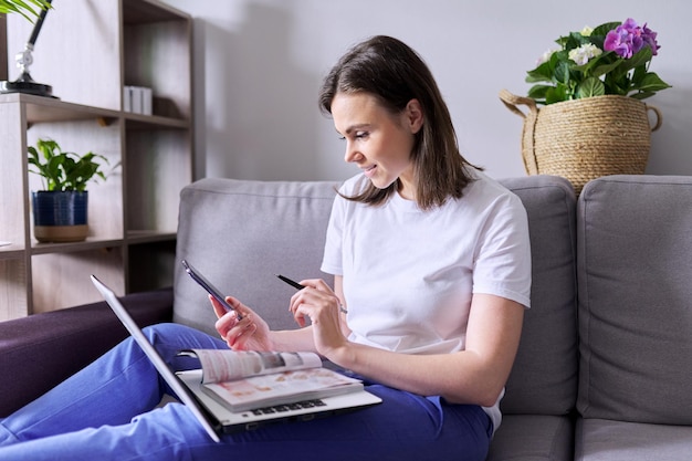 Une jeune femme achète des produits, des cosmétiques, des vêtements en ligne. Entre les mains d'un smartphone pour ordinateur portable à catalogue papier, la femme fait des achats à la maison, assise sur le canapé