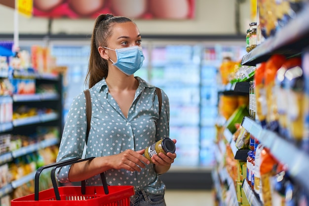 Jeune femme acheteuse portant un masque de protection médicale parmi les étagères choisit des produits alimentaires dans une épicerie