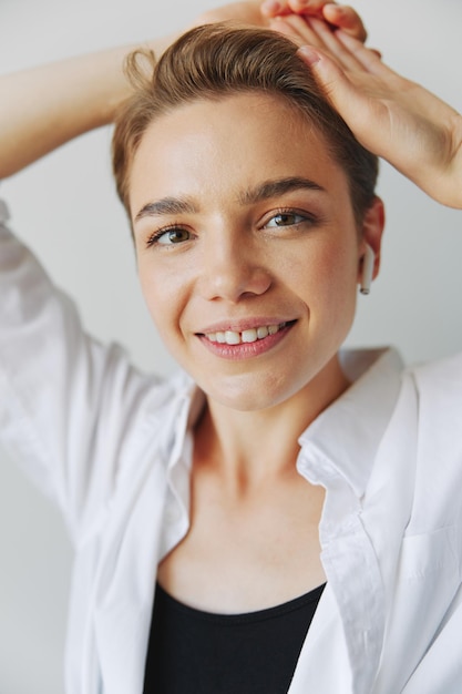 Jeune femme adolescente écoutant de la musique avec des écouteurs infertiles et dansant à la maison souriant avec des dents avec une coupe de cheveux courte dans une chemise blanche sur fond blanc Fille pose naturelle sans filtres