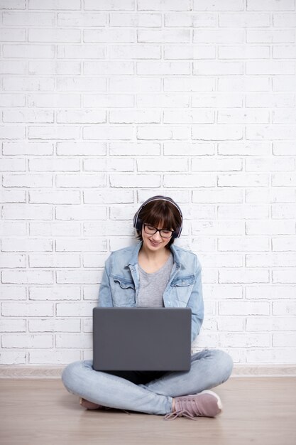 Jeune femme ou adolescente utilisant un ordinateur portable et écoutant de la musique dans des écouteurs sur fond de mur de briques blanches avec espace pour copie