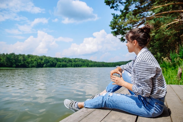 Jeune femme adulte assise sur un quai en bois buvant du café et regardant le lac