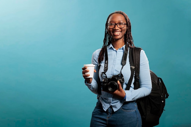 Jeune femme adulte avec passe-temps de photographie souriant chaleureusement à la caméra. Heureux amateur de photographie positive ayant un appareil photo DSLR et une tasse de café en se tenant debout sur fond bleu.