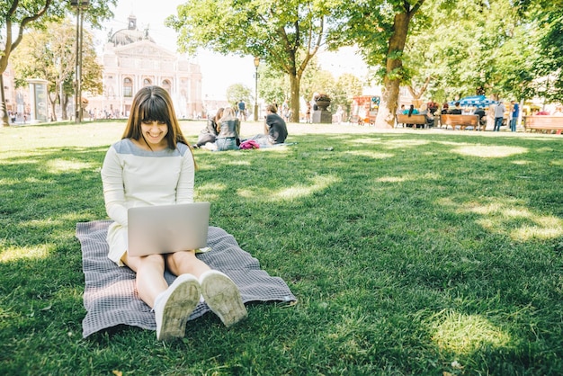 Jeune femme adulte portant avec un ordinateur portable dans le parc de la ville sur l'herbe verte