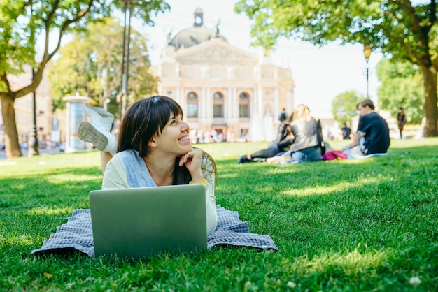 Jeune femme adulte portant avec un ordinateur portable dans le parc de la ville sur l'herbe verte