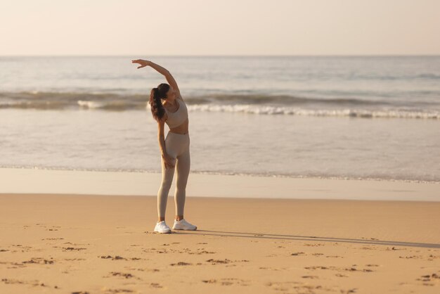 Une jeune femme adulte en tenue d'entraînement s'étire les mains et le corps sur la plage le matin.