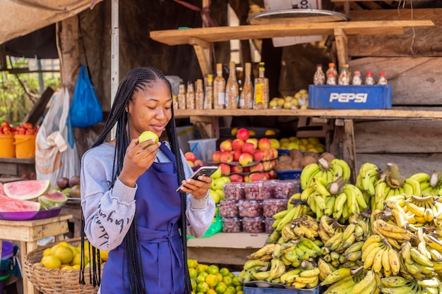 Jeune femme d'affaires africaine noire dans un marché local mangeant une pomme naviguant en ligne à l'aide d'un smartphone vérifiant la lecture des nouvelles en ligne