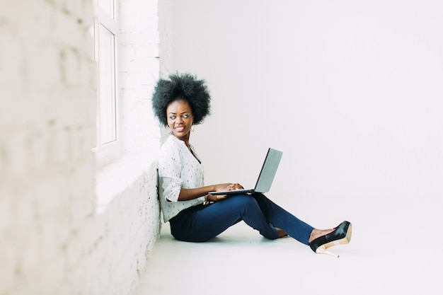 Jeune femme d'affaires afro-américaine à l'aide de l'ordinateur portable, assis sur le sol près d'une grande fenêtre