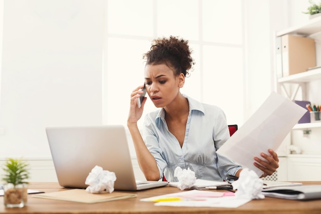 Jeune femme d'affaires afro-américaine sérieuse parlant par téléphone avec des papiers, assise sur un lieu de travail de bureau moderne. Conseil aux entreprises.