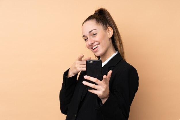 Jeune femme d'affaires à l'aide de téléphone portable sur le mur isolé pointe le doigt vers vous avec une expression confiante