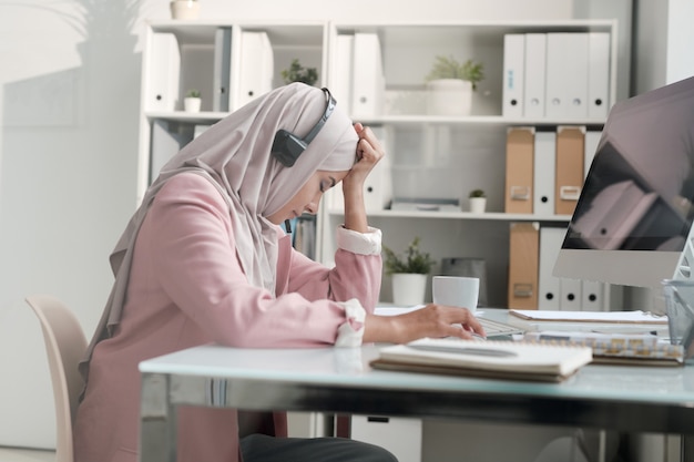 Jeune femme d'affaires arabe en hijab fatigué de travailler dans un centre d'appels assis au bureau et se penchant la tête sur place