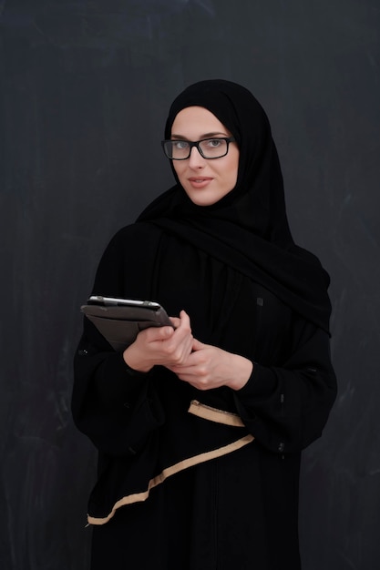 Jeune femme d'affaires arabe en vêtements traditionnels ou abaya et lunettes tenant une tablette devant un tableau noir représentant la mode et la technologie de l'islam moderne