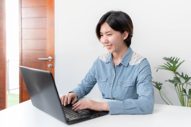 Jeune femme d'affaires asiatique assise à la maison travaillant sur son bureau et souriant joyeusement à son travail