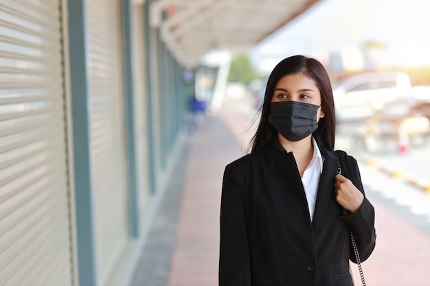 Jeune femme d'affaires asiatique en costume noir d'affaires avec masque de protection pour les soins de santé marchant sur la rue publique en plein air et à la recherche. Nouveau concept de distanciation normale et sociale