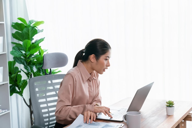 Jeune femme d'affaires asiatique enthousiaste au bureau moderne utilisant un ordinateur portable pour travailler et écrire des notes Dame de bureau diligente et attrayante travaillant sur un ordinateur portable dans son espace de travail de bureau