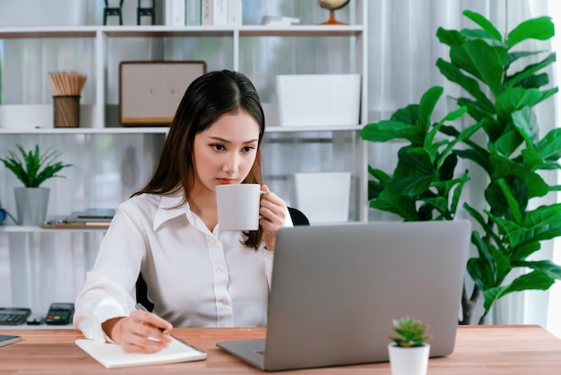 Jeune femme d'affaires asiatique enthousiaste au bureau moderne utilisant un ordinateur portable pour travailler avec une tasse de café Dame de bureau diligente et attrayante travaillant sur un ordinateur portable dans son espace de travail de bureau