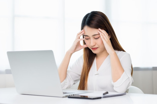 Jeune femme d'affaires asiatique main touchant la tête, elle se sent déprimée, stress, maux de tête, être fatiguée de travailler avec un ordinateur portable pendant longtemps, elle a le syndrome du bureau des symptômes.