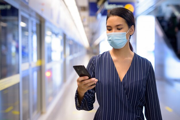 Jeune femme d'affaires asiatique avec masque à l'aide de téléphone en attendant à la station de métro