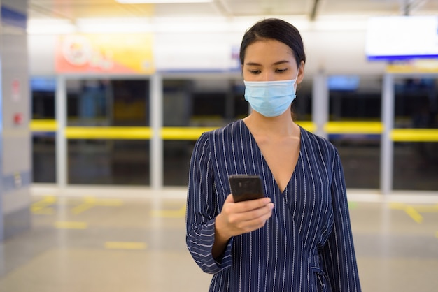 Jeune femme d'affaires asiatique avec masque à l'aide de téléphone à la station de métro