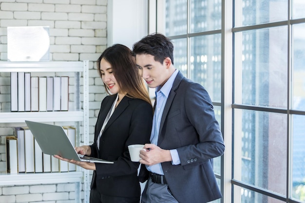 Jeune femme d'affaires asiatique et partenaires d'affaires tout en travaillant avec un ordinateur portable et un homme d'affaires tenant une tablette en arrière-plan de bureauCouple de travail