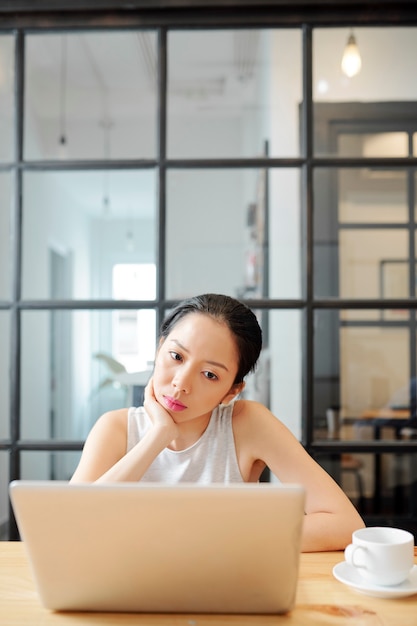 Jeune femme d'affaires asiatique pensive regardant une présentation ou lisant un rapport sur un écran d'ordinateur portable lorsqu'elle est assise à une table basse avec une tasse de café