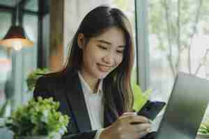 Photo une jeune femme d'affaires asiatique souriante qui utilise et regarde son téléphone portable alors qu'elle travaille sur un ordinateur portable