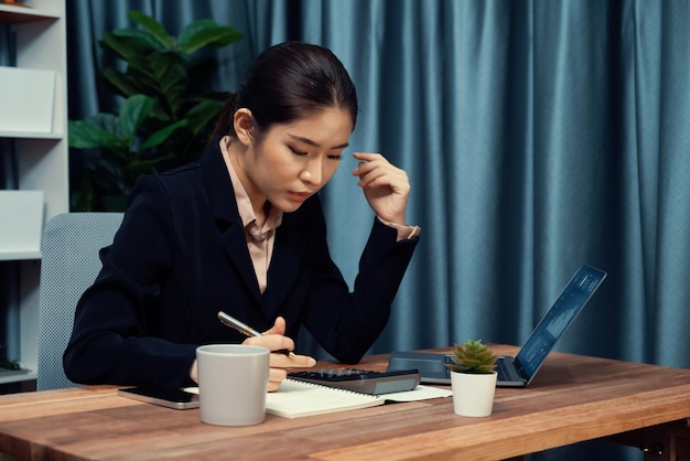 Une jeune femme d'affaires asiatique travaille diligemment et prend des notes dans son bureau moderne, démontrant son professionnalisme en tant que femme de bureau attrayante et diligente.