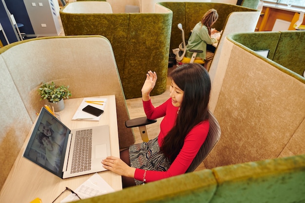 Jeune femme d'affaires asiatique en vêtements de sport intelligents et masque de protection agitant la main tout en regardant un couple heureux sur un écran d'ordinateur portable