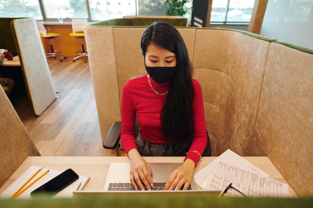 Jeune femme d'affaires asiatique vêtue de vêtements décontractés intelligents et d'un masque de protection regardant un écran d'ordinateur portable assis à son bureau et en réseau