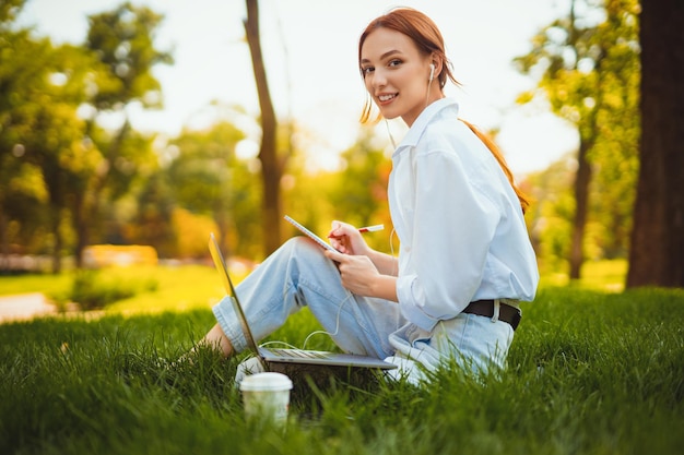 Une jeune femme d'affaires assise sur l'herbe dans un parc utilise un ordinateur portable pour travailler prend des notes à l'extérieur du bureau