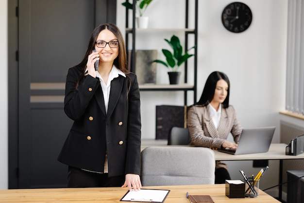 Une jeune femme d'affaires attrayante portant une veste parle au téléphone portable alors qu'elle est assise sur un bureau et utilise un bloc-notes près de son collègue au travail au bureau