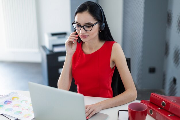 Jeune femme d'affaires attrayante en robe rouge et lunettes s'asseoir à la table et travaille avec un ordinateur portable