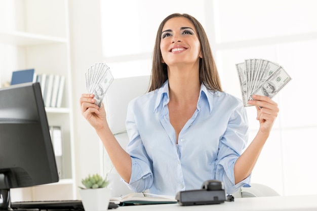 Jeune femme d'affaires au bureau, assise à un bureau, tenant de l'argent et avec une expression de bonheur sur son visage en levant.