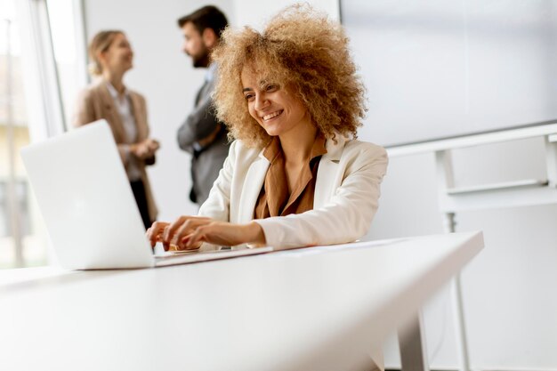 Jeune femme d'affaires aux cheveux bouclés utilisant un ordinateur portable au bureau avec des jeunes travaille derrière elle