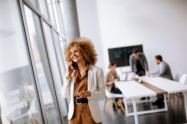 Jeune femme d'affaires aux cheveux bouclés utilisant un téléphone portable au bureau avec des jeunes travaille derrière elle