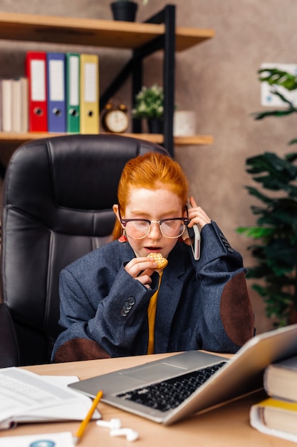 Jeune femme d'affaires. Belle fille positive mangeant un cookie tout en passant un appel téléphonique