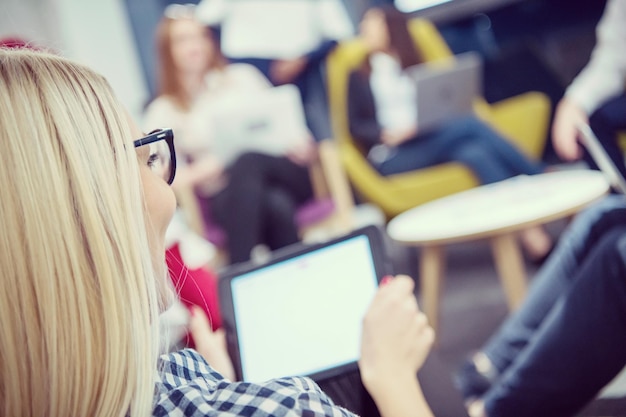 jeune femme d'affaires blonde travaillant en ligne à l'aide d'une tablette numérique tout en étant assise au bureau de démarrage moderne avec une équipe commerciale multiethnique en arrière-plan