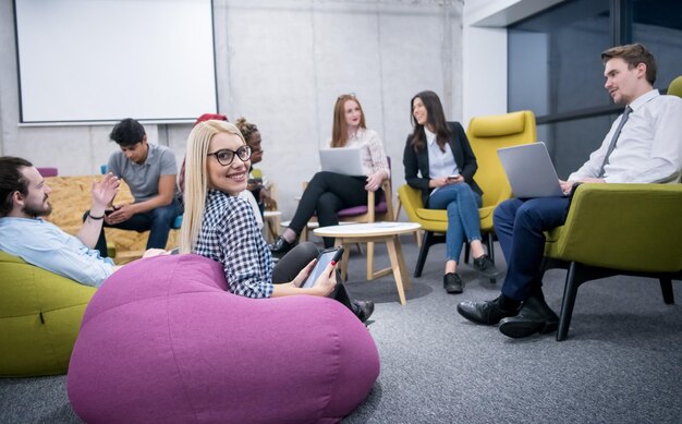 jeune femme d'affaires blonde travaillant en ligne à l'aide d'une tablette numérique tout en étant assise au bureau de démarrage moderne avec une équipe commerciale multiethnique en arrière-plan