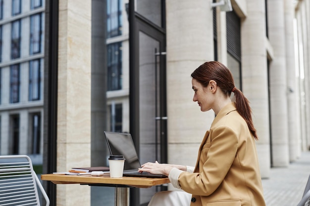 Une jeune femme d'affaires brune en tenue de cérémonie beige utilisant un ordinateur portable dans un café de rue