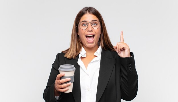 Jeune femme d'affaires avec un café se sentant comme un génie heureux et excité après avoir réalisé une idée, levant joyeusement le doigt, eureka!