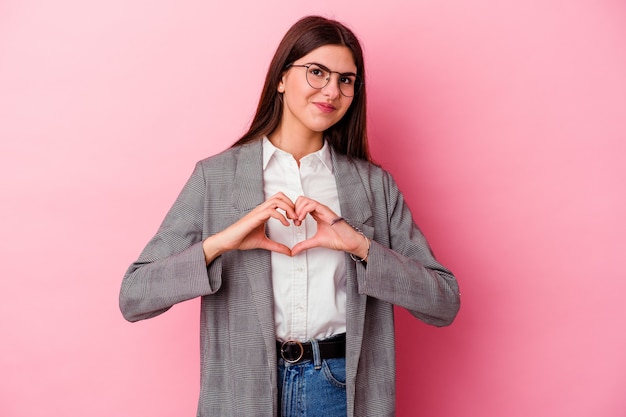 Jeune femme d'affaires caucasienne isolée sur fond rose souriant et montrant une forme de coeur avec les mains.