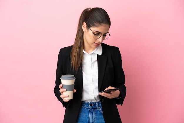 Jeune femme d'affaires caucasienne isolée sur fond rose tenant du café à emporter et un mobile