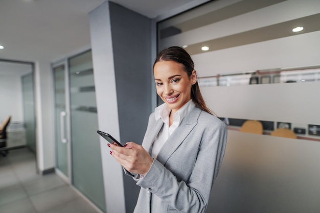 Jeune femme d'affaires caucasienne magnifique réussie souriante debout dans le hall de l'entreprise, tenant un téléphone intelligent et prenant une pause.