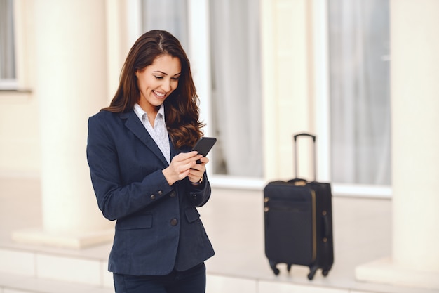 Jeune femme d'affaires caucasienne souriante habillée décontractée intelligente à l'aide d'un téléphone intelligent pour écrire un message tout en se tenant à l'extérieur. En arrière-plan des bagages. Concept de voyage d'affaires.