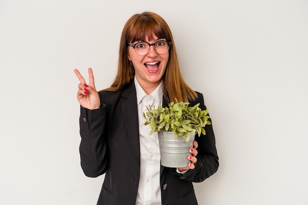 Jeune femme d'affaires caucasienne tenant une plante isolée sur fond blanc joyeuse et insouciante montrant un symbole de paix avec les doigts.