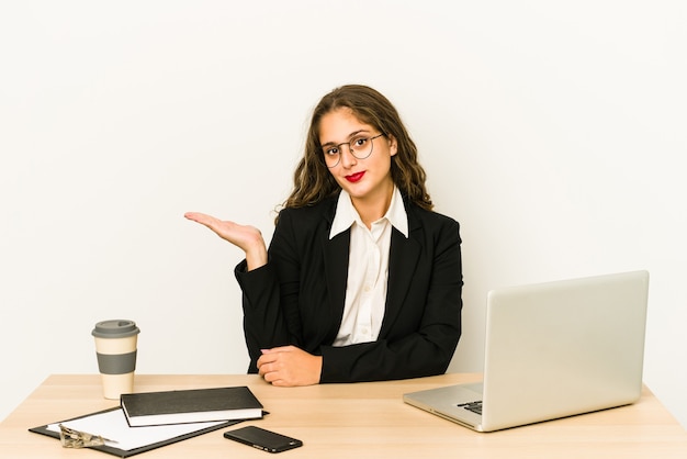 Jeune femme d'affaires caucasienne travaillant sur son bureau montrant un espace de copie sur une paume et tenant une autre main sur la taille.